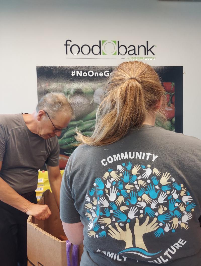a woman, facing away, and a man place food into a large cardboard box